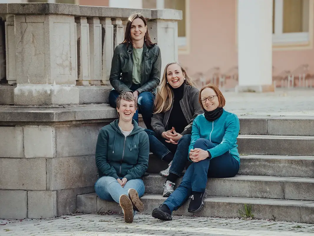 Vier Frauen des Teams von „wir unterwegs“ sitzen gemeinsam auf einer Treppe. Sie lächeln und blicken in die Kamera. Das Bild strahlt Teamgeist, Offenheit und Zusammenhalt aus.