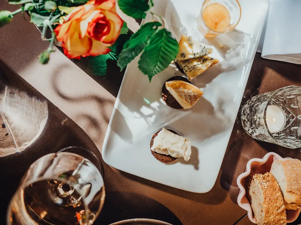 Nahaufnahme eines gedeckten Tisches mit Käseauswahl, Brot, Wein und einer Blume – stimmungsvolle Szene eines kulinarischen Highlights während einer intensiven Gruppenreise oder eines Teamevents.
