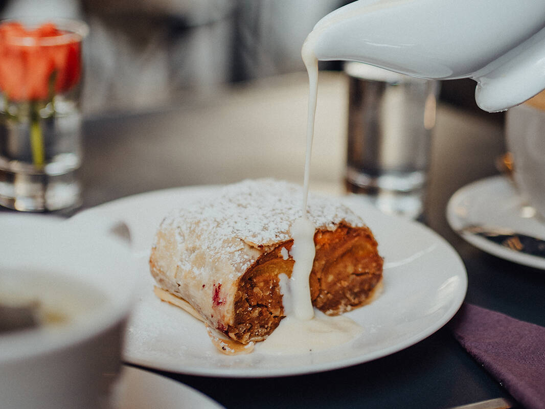 Nahaufnahme eines Strudels im Strudel Café Kröll in Innsbruck. Auf einem Teller wird Vanillesoße über den warmen Strudel gegossen, im Hintergrund Caféatmosphäre.