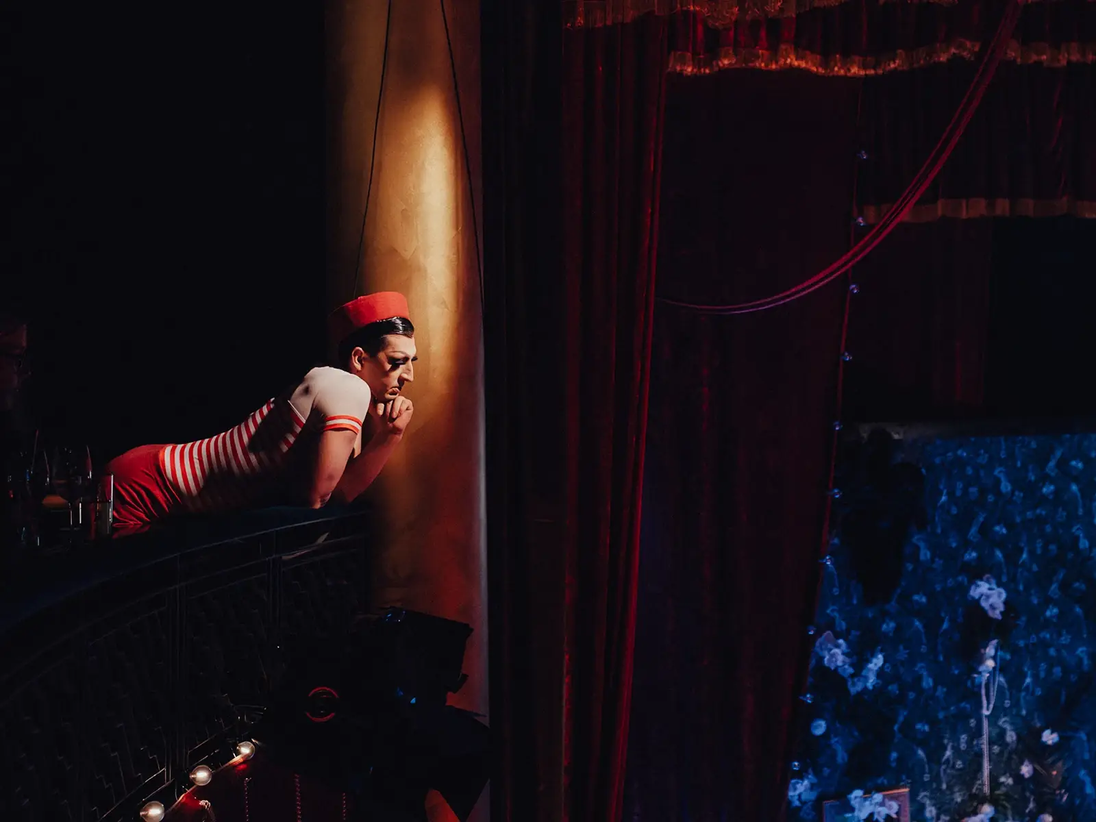 Künstler in rot weißem Kostüm lehnt sich von einer Galerie über das Geländer und blickt zur Bühne im Krystallpalast Varieté Leipzig, roter Vorhang und Theaterlicht im Hintergrund.