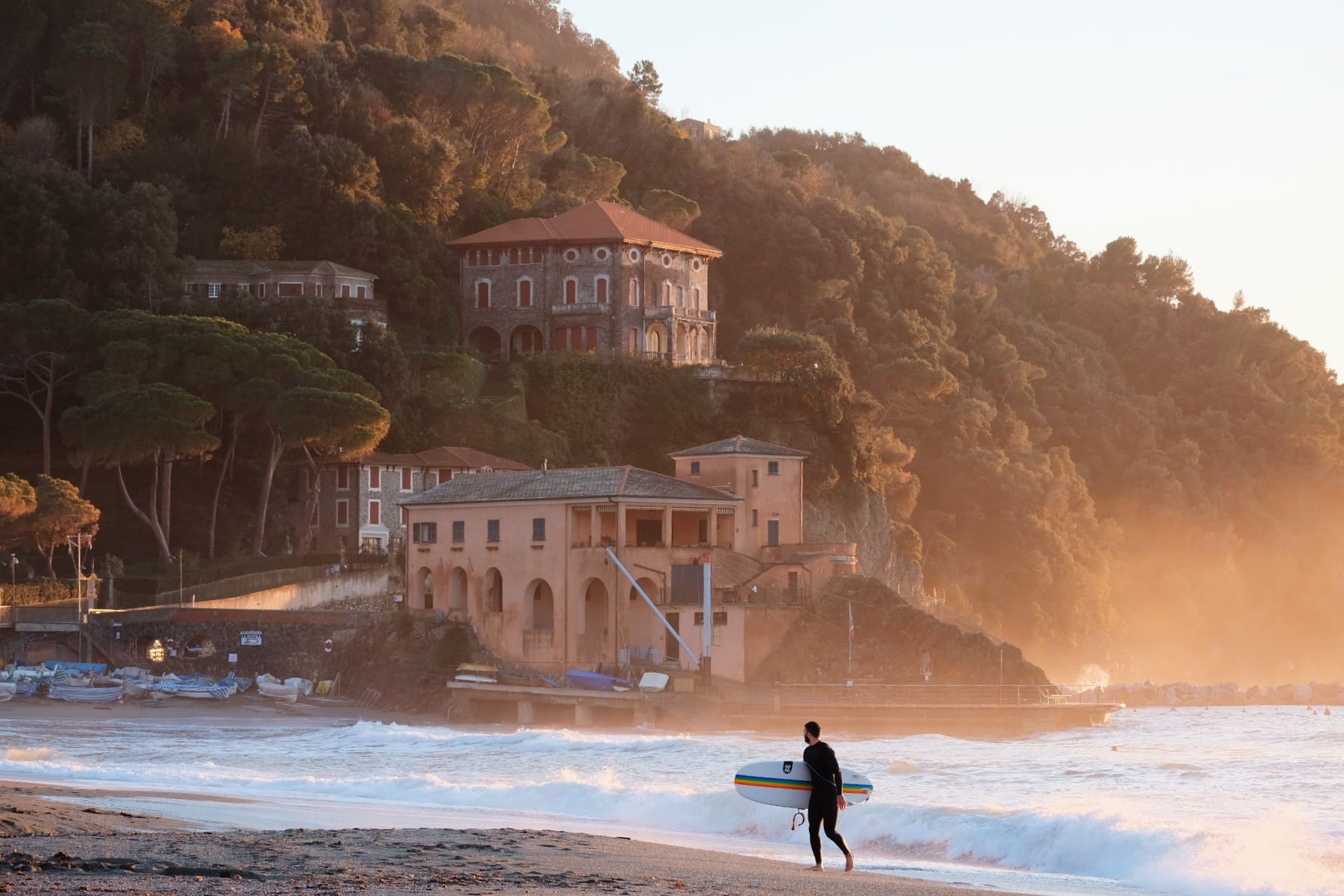 Levanto in Ligurien bei Sonnenuntergang mit historischem Küstengebäude, bewaldeten Hügeln und Surfer am Strand.