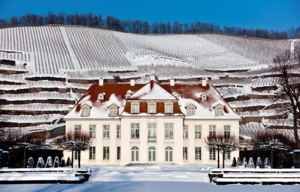 Das schneebedeckte Schloss Wackerbarth in Radebeul im altbarocken Baustil vor dem verschneiten Weinberg des anliegenden Weinguts.